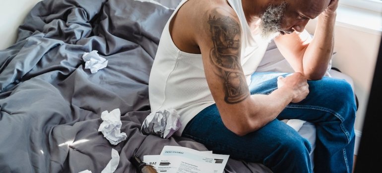 Depressed man sitting on his bed next to a pile of overdue bills and an empty glass bottle