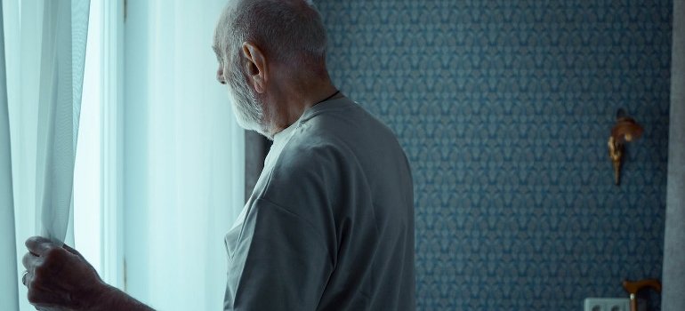 An elderly man is standing near the window while thinking of veterans' substance abuse stats for NJ and NY.