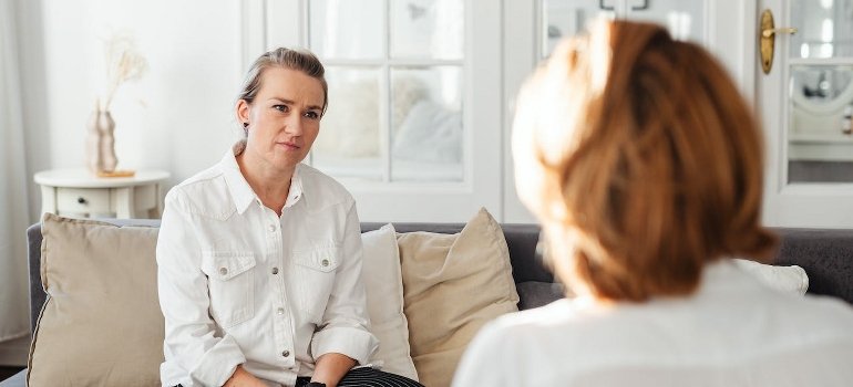 The two women are sitting and talking at home about family members of addicts.