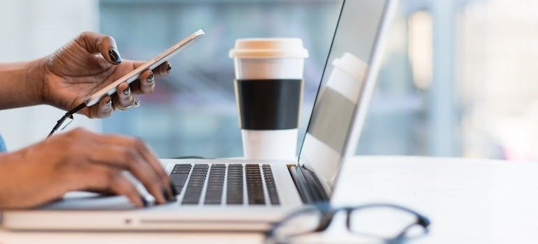 A person holding a phone while typing on a laptop, doing research on common rehab and addiction myths and How Binghamton NY Residents Can Access Top-Notch Rehab in PA