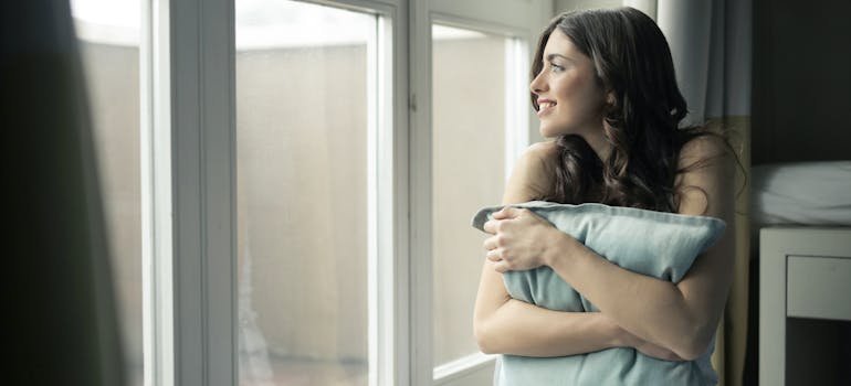 woman smiling and looking out the window while holding her pillow