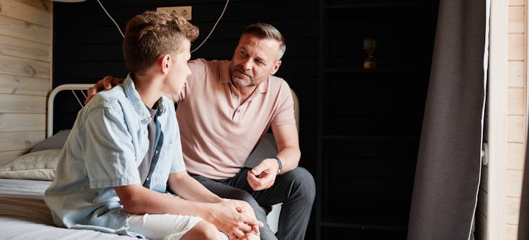 a father and a son sitting on a bed and talking