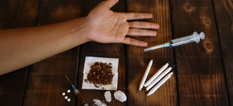 Black tar heroine over a small piece of paper alongside crystallized drugs with a syringe and the had of a person