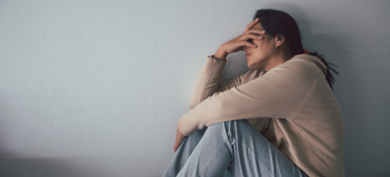 A woman against a white wall dressed in sweats and a sweater covering her head with one of her hands, dreading her past actions