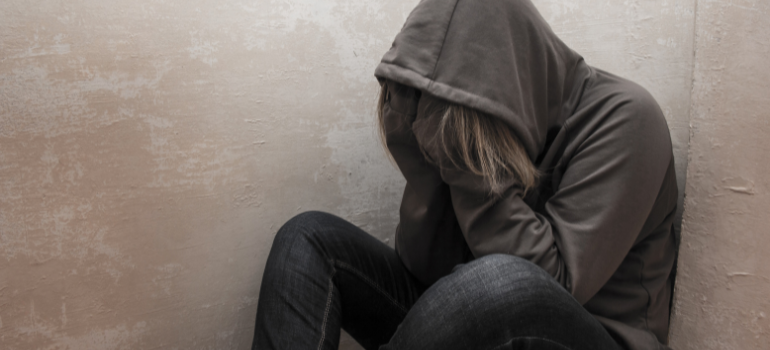 A long-haired person with a dark gray hoodie covering their face and jeans looks distressed against a wall thinking about black tar heroin