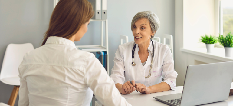 A young woman with long hair and a white button-up shirt shows her back as she talks to a medical doctor talking about black tar heroin