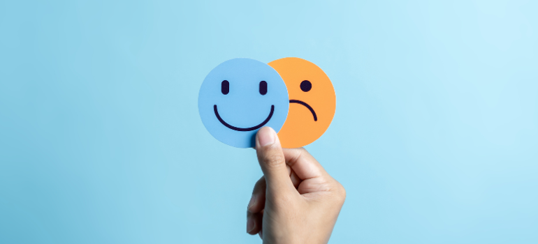 A human hand holds two paper cutouts, one with a smiley face and one with a sad face, against a bright blue wall.