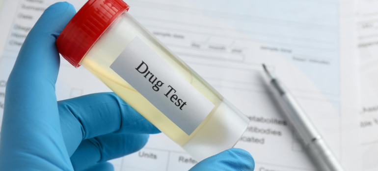 A gloved hand holding a vial with a yellow liquid, some forms and a pen in the background