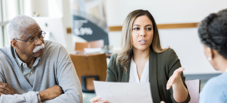 A woman dressed in a business suit explains something written on a sheet of paper she is holding in her hand to an older man and another woman