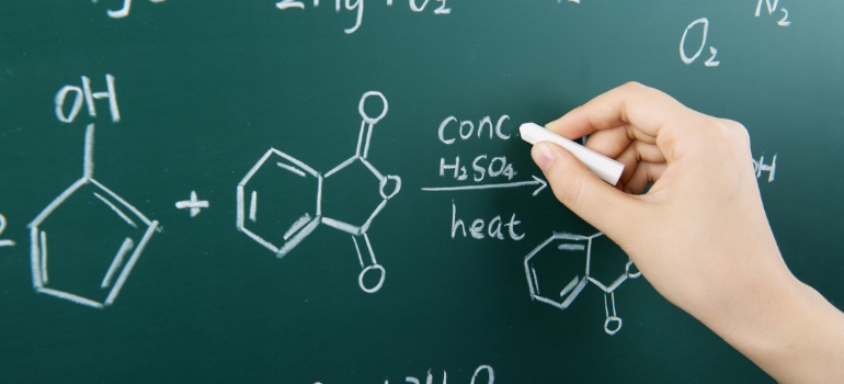 The hand of a person holding chalk while solving a chemistry problem on the board. 
