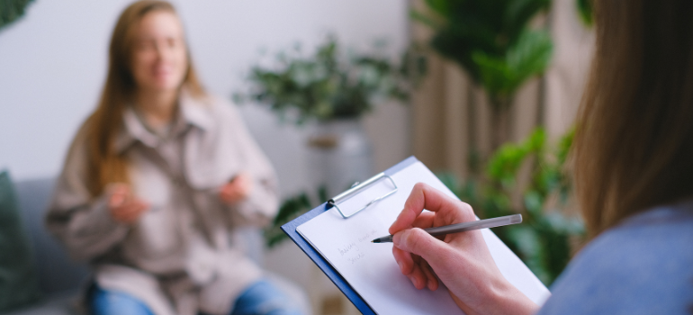 A therapist takes notes while her patient, blurred in the background talks about why people use cocaine when drinking alcohol