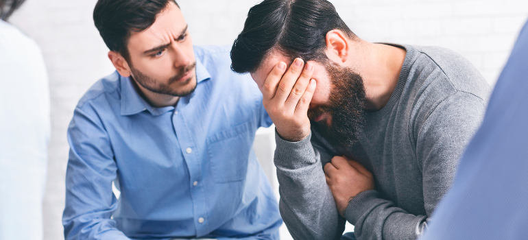 A young therapist comforts his patient, who has his hands on his face and looks distressed after talking about exploring the dangers and realities of bath salts