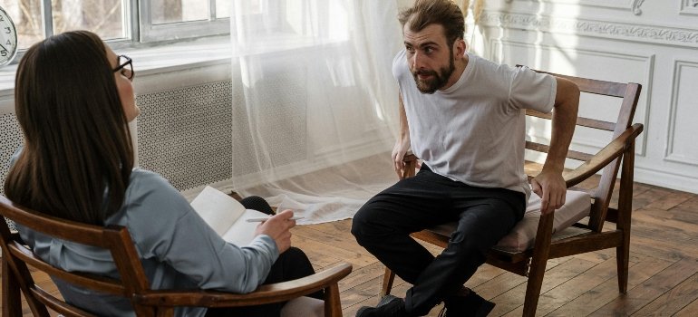 A man in a white T-shirt is sitting on a wooden chair and talking about drinking problems with his psychologist.