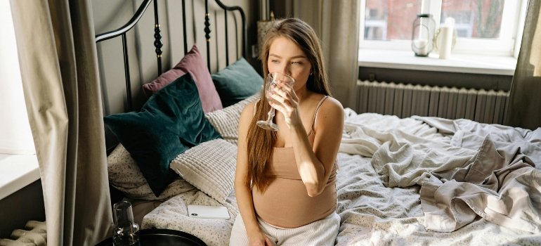 A pregnant woman is sitting on the bed drinking water from a glass.