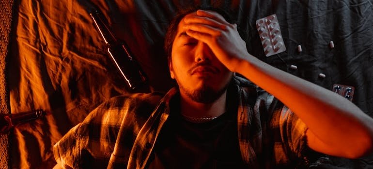 Man lying on the bed with a beer bottle and blisters of pills around him.