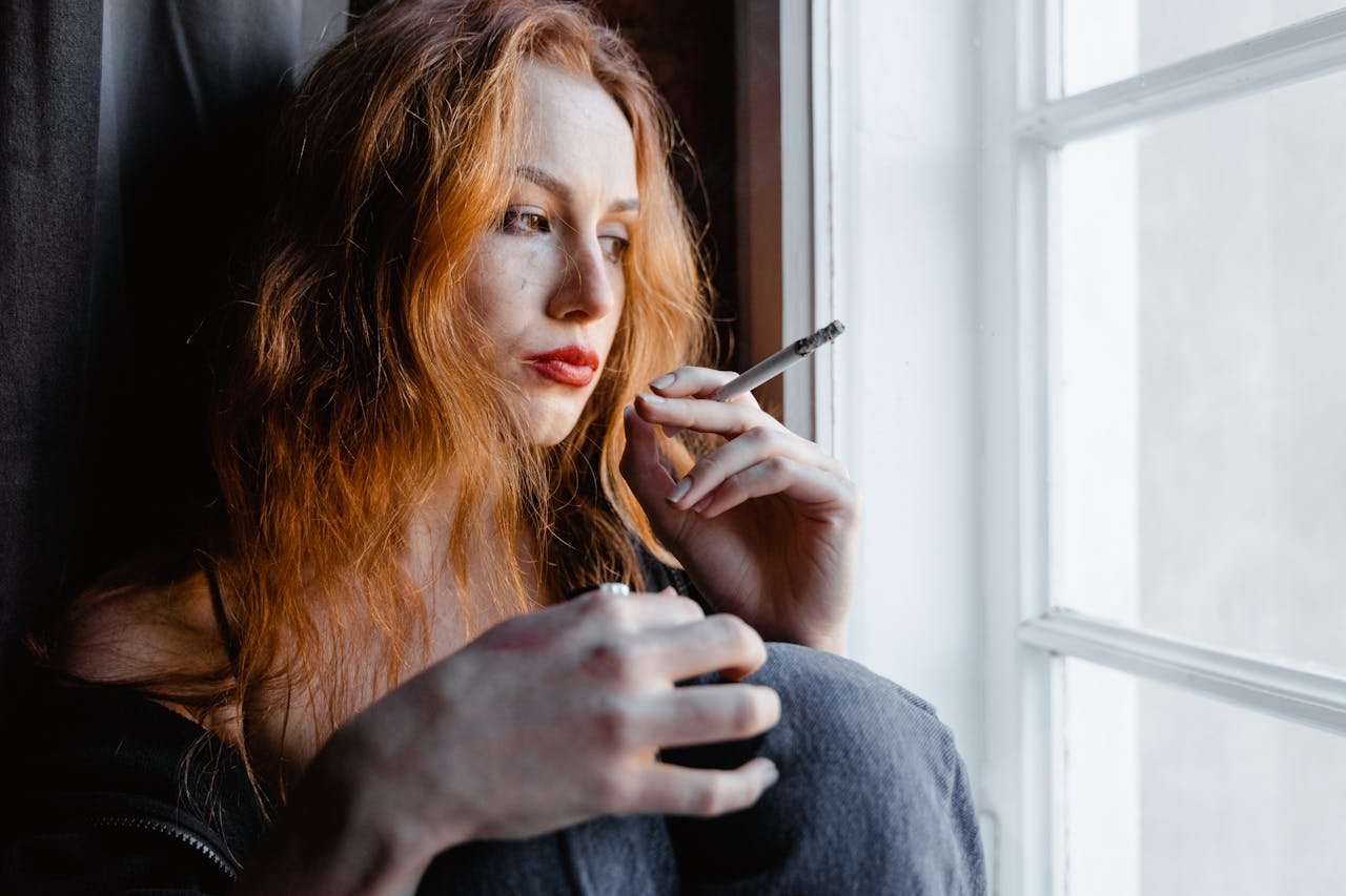 Woman looking out of a window and smoking a cigarette while struggling with depression and addiction.
