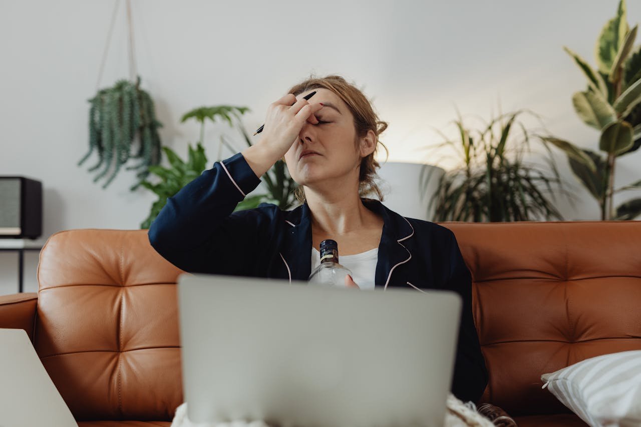Stressed out woman holding a bottle of alcohol while working on her laptop.
